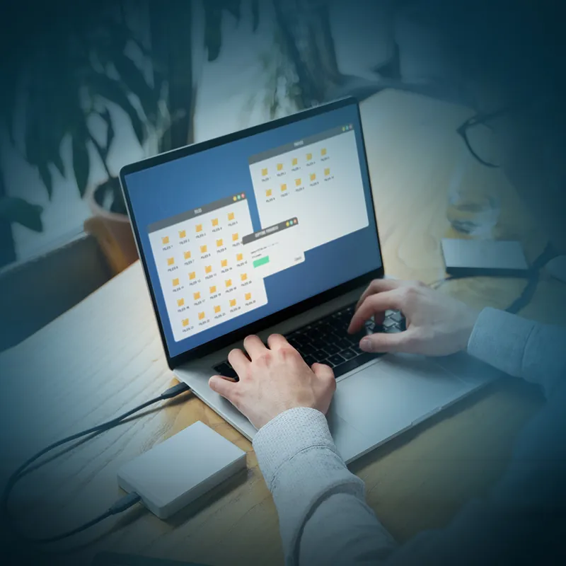 يدان تكتبان على حاسب محمول تعرض شاشته نوافذ مجلدات ونافذة عملية جارية، وقرص خارجي على الطاولة